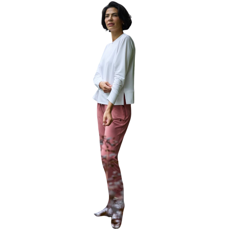 A person with long black hair is standing against a plain white background, wearing a Cozy Earth Women's StudioLite Crewneck in light green and dark green pants. They are looking down with a gentle smile, gently touching their neck with one hand. |Color:Haze