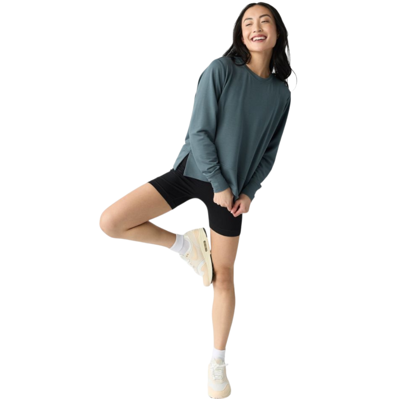 A person with long black hair is standing against a plain white background, wearing a Cozy Earth Women's StudioLite Crewneck in light green and dark green pants. They are looking down with a gentle smile, gently touching their neck with one hand. |Color:Haze