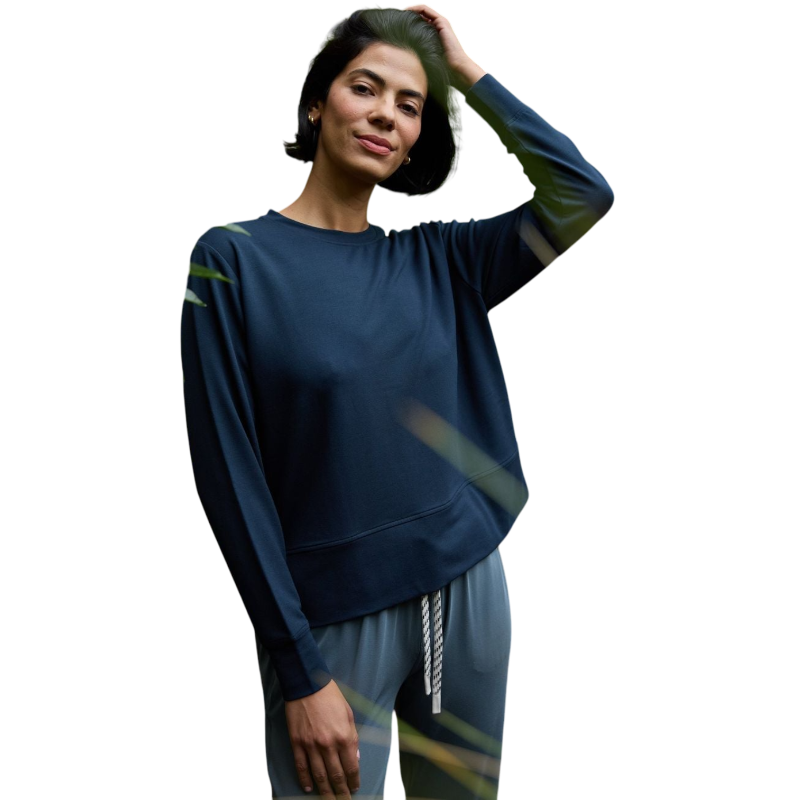 A person with long black hair is standing against a plain white background, wearing a Cozy Earth Women's StudioLite Crewneck in light green and dark green pants. They are looking down with a gentle smile, gently touching their neck with one hand. |Color:Haze