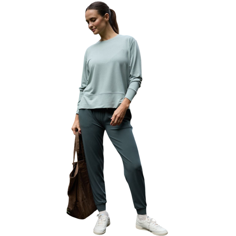 A person with long black hair is standing against a plain white background, wearing a Cozy Earth Women's StudioLite Crewneck in light green and dark green pants. They are looking down with a gentle smile, gently touching their neck with one hand. |Color:Haze