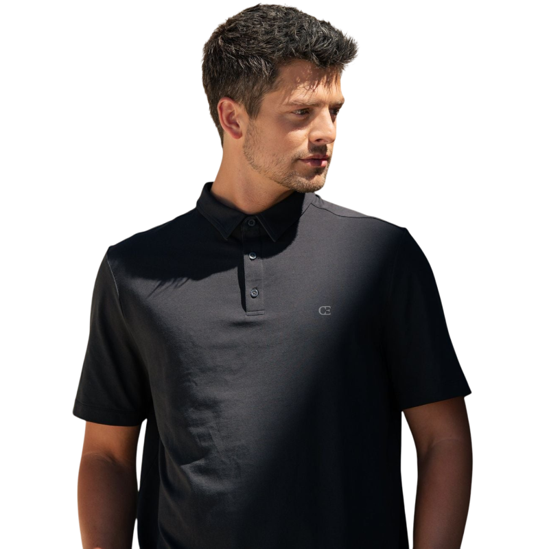 A person with short, light brown hair is wearing the Men's Everyday Polo by Cozy Earth in a light gray shade, paired with light beige pants. They are standing and slightly looking over their left shoulder with a neutral expression against a plain white background. |Color:Stone