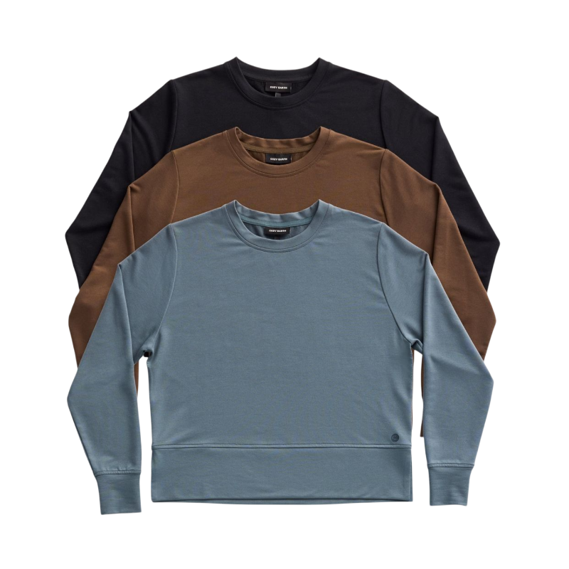 A person with long black hair is standing against a plain white background, wearing a Cozy Earth Women's StudioLite Crewneck in light green and dark green pants. They are looking down with a gentle smile, gently touching their neck with one hand. |Color:Haze