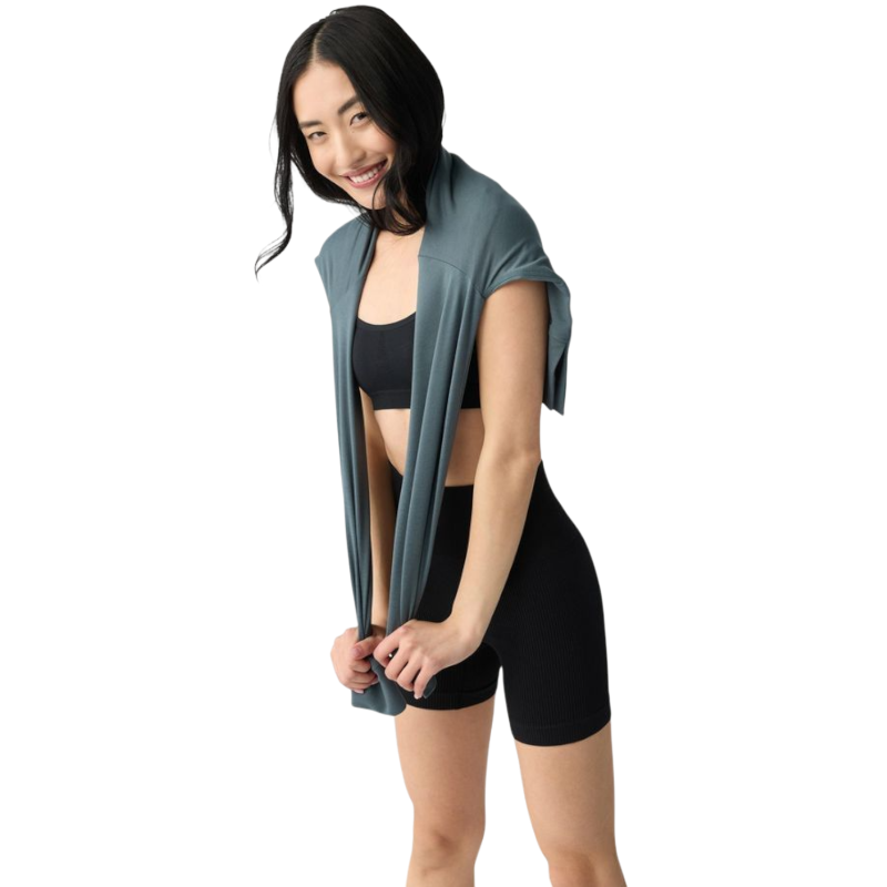 A person with long black hair is standing against a plain white background, wearing a Cozy Earth Women's StudioLite Crewneck in light green and dark green pants. They are looking down with a gentle smile, gently touching their neck with one hand. |Color:Haze