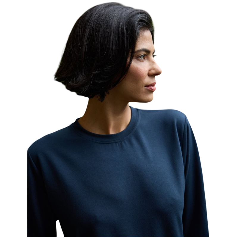 A person with long black hair is standing against a plain white background, wearing a Cozy Earth Women's StudioLite Crewneck in light green and dark green pants. They are looking down with a gentle smile, gently touching their neck with one hand. |Color:Haze
