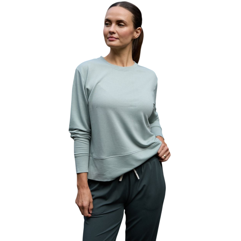 A person with long black hair is standing against a plain white background, wearing a Cozy Earth Women's StudioLite Crewneck in light green and dark green pants. They are looking down with a gentle smile, gently touching their neck with one hand. |Color:Haze