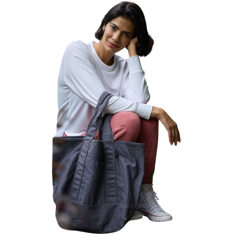 A person with long black hair is standing against a plain white background, wearing a Cozy Earth Women's StudioLite Crewneck in light green and dark green pants. They are looking down with a gentle smile, gently touching their neck with one hand. |Color:Haze