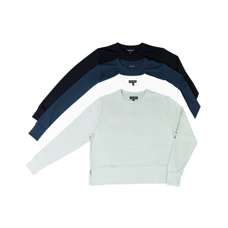 A person with long black hair is standing against a plain white background, wearing a Cozy Earth Women's StudioLite Crewneck in light green and dark green pants. They are looking down with a gentle smile, gently touching their neck with one hand. |Color:Haze