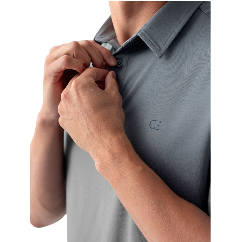 A person with short, light brown hair is wearing the Men's Everyday Polo by Cozy Earth in a light gray shade, paired with light beige pants. They are standing and slightly looking over their left shoulder with a neutral expression against a plain white background. |Color:Stone