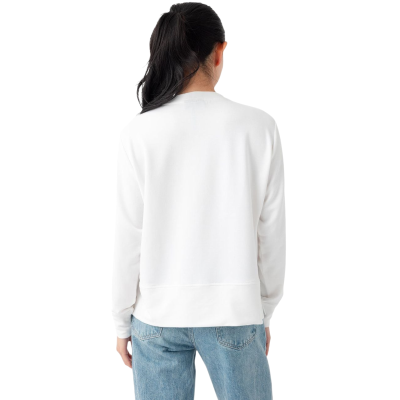 A person with long black hair is standing against a plain white background, wearing a Cozy Earth Women's StudioLite Crewneck in light green and dark green pants. They are looking down with a gentle smile, gently touching their neck with one hand. |Color:Haze