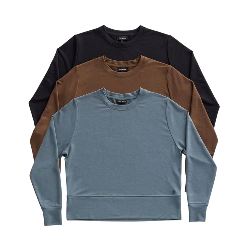 A person with long black hair is standing against a plain white background, wearing a Cozy Earth Women's StudioLite Crewneck in light green and dark green pants. They are looking down with a gentle smile, gently touching their neck with one hand. |Color:Haze