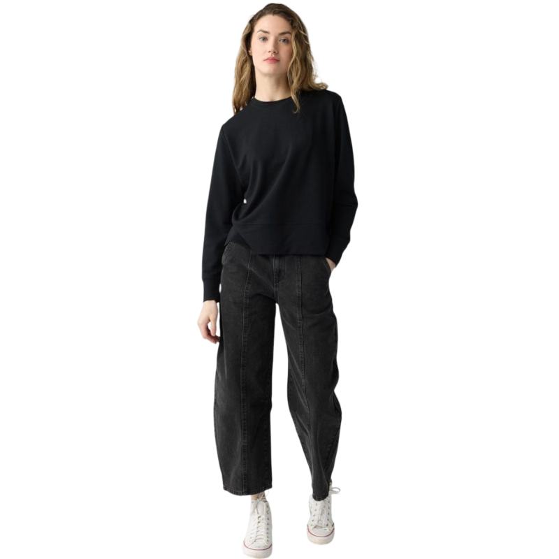 A person with long black hair is standing against a plain white background, wearing a Cozy Earth Women's StudioLite Crewneck in light green and dark green pants. They are looking down with a gentle smile, gently touching their neck with one hand. |Color:Haze