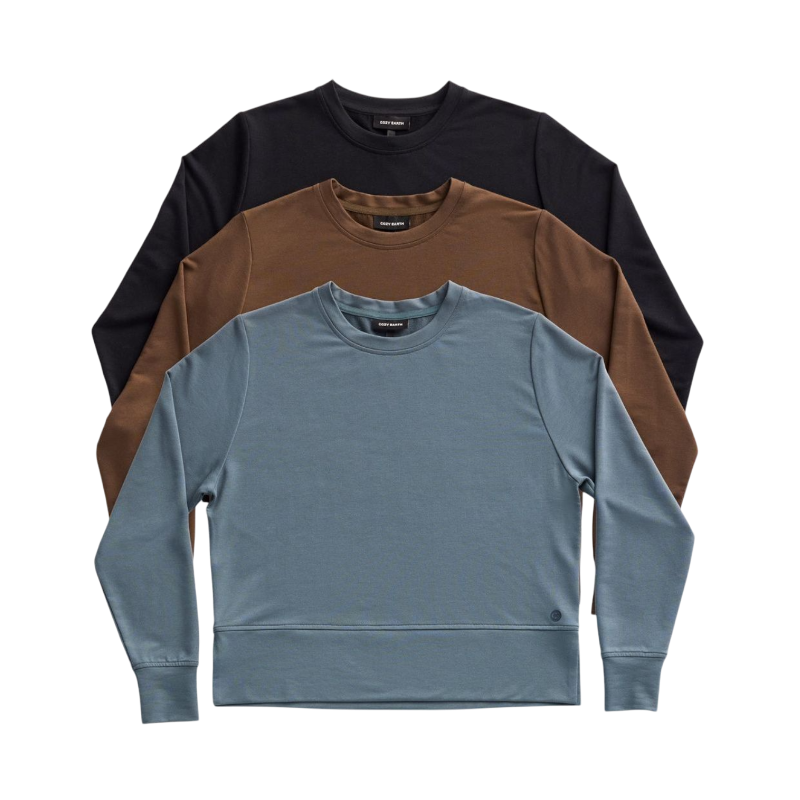 A person with long black hair is standing against a plain white background, wearing a Cozy Earth Women's StudioLite Crewneck in light green and dark green pants. They are looking down with a gentle smile, gently touching their neck with one hand. |Color:Haze