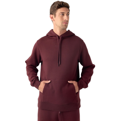 A man is wearing a maroon Men's CityScape Hoodie by Cozy Earth, looking slightly to the left. He has short brown hair and his hands are tucked into the front pocket of the hoodie. The background is plain white. |Color:Burgundy