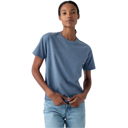 A person with long hair pulled back is wearing Cozy Earth's Women's All Day Tee, a blue short-sleeve t-shirt, along with light blue jeans. They are standing against a plain white background with a neutral expression, holding their hands near the waistband of their jeans. |Color:Deep Blue Heather