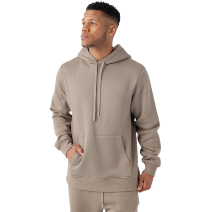 A man stands against a plain white background, wearing Cozy Earth's Men's CityScape Hoodie in a taupe color with a front pocket and matching joggers. He has short hair and is looking to his left with one hand partially in his pocket. |Color:Ebony