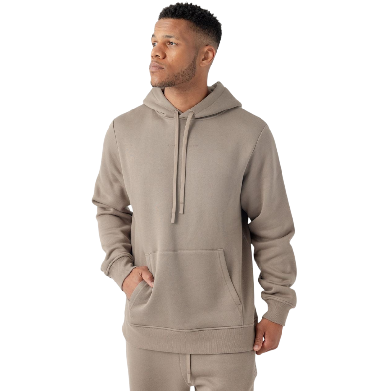 A man stands against a plain white background, wearing Cozy Earth's Men's CityScape Hoodie in a taupe color with a front pocket and matching joggers. He has short hair and is looking to his left with one hand partially in his pocket. |Color:Ebony