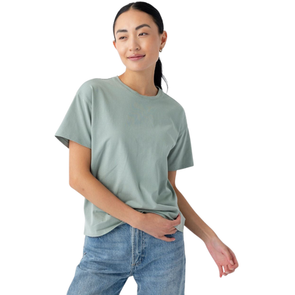 A person with long dark hair tied back is wearing the Women's All Day Tee in light green and blue jeans. They are standing against a plain white background, holding part of the t-shirt with one hand and smiling slightly. The t-shirt is by Cozy Earth. |Color:Haze
