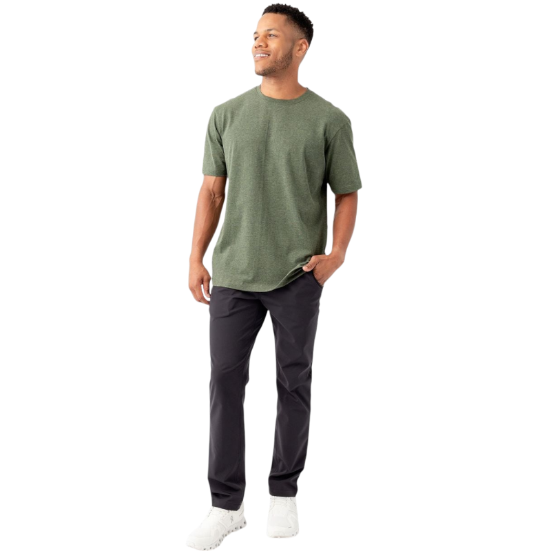 A man is smiling while standing against a plain white background, wearing a Cozy Earth Men's All Day Tee in green, paired with black pants and white sneakers. One of his hands is in his pocket while the other rests relaxed at his side. |Color:Olive Heather