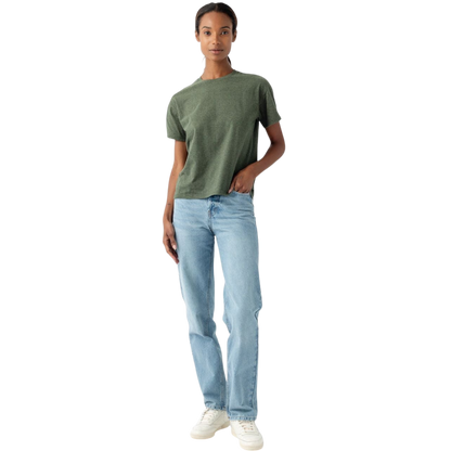 Against a plain white background, a person dressed in Cozy Earth's Women's All Day Tee, which is short-sleeved and green, pairs it with light blue jeans and white sneakers. One hand hangs relaxed by their side while the other rests casually in their front pocket. Their expression is calm and neutral.