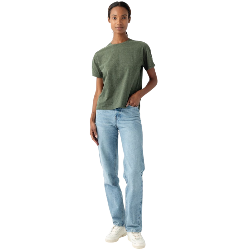 Against a plain white background, a person dressed in Cozy Earth's Women's All Day Tee, which is short-sleeved and green, pairs it with light blue jeans and white sneakers. One hand hangs relaxed by their side while the other rests casually in their front pocket. Their expression is calm and neutral.