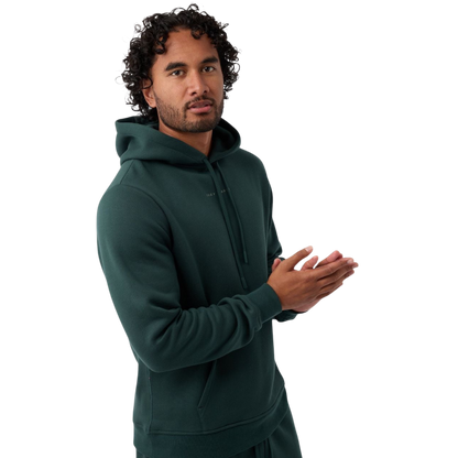 A person with curly hair is wearing a Cozy Earth Men's CityScape Hoodie in dark green and standing against a plain white background, with their hands together. |Color:Spruce