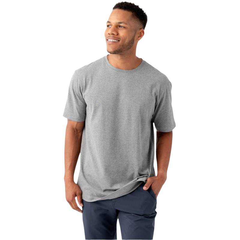 A man stands against a white background, smiling and looking to the side. He is dressed casually in a plain gray Men's All Day Tee by Cozy Earth and dark blue pants. One hand is in his pocket, while the other hangs by his side. |Color:Stone Heather