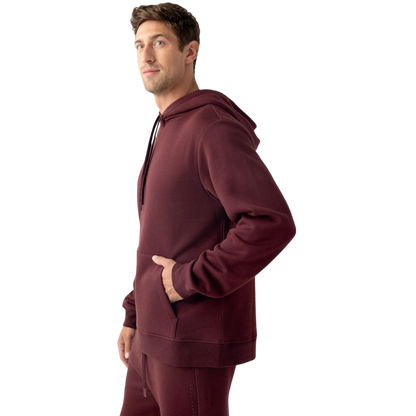 A person with curly hair is wearing a Cozy Earth Men's CityScape Hoodie in dark green and standing against a plain white background, with their hands together. |Color:Spruce