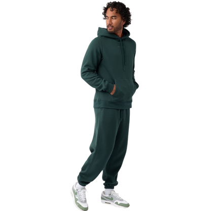 A person with curly hair is wearing a Cozy Earth Men's CityScape Hoodie in dark green and standing against a plain white background, with their hands together. |Color:Spruce