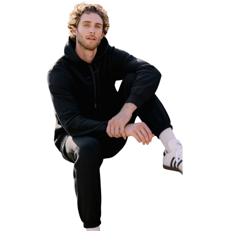 A person with curly hair is wearing a Cozy Earth Men's CityScape Hoodie in dark green and standing against a plain white background, with their hands together. |Color:Spruce
