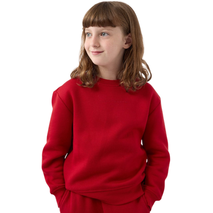 A young person with brown hair stands wearing a bright red Kid's CityScape Crewneck from Cozy Earth and pants against a plain white background, looking to the side and smiling slightly, with their hands tucked into their pockets. |Color:Crimson