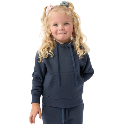 A young child with long blonde hair smiles while wearing a navy blue Kid's CityScape Hoodie by Cozy Earth and matching pants. She has two hair ties, one pink and one blue. The background is plain white. |Color:Eclipse