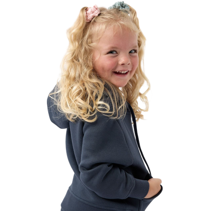 A young child with long blonde hair smiles while wearing a navy blue Kid's CityScape Hoodie by Cozy Earth and matching pants. She has two hair ties, one pink and one blue. The background is plain white. |Color:Eclipse