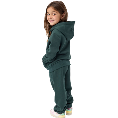 A young child with long blonde hair smiles while wearing a navy blue Kid's CityScape Hoodie by Cozy Earth and matching pants. She has two hair ties, one pink and one blue. The background is plain white. |Color:Eclipse