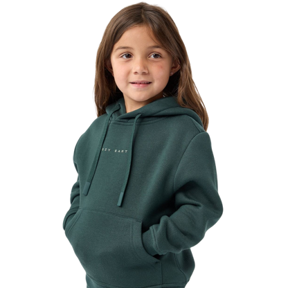 A young girl with long brown hair is wearing a Kid's CityScape Hoodie by Cozy Earth, featuring the brand logo. She has her hands in the hoodie pockets and is smiling slightly, standing against a plain white background. |Color:Spruce