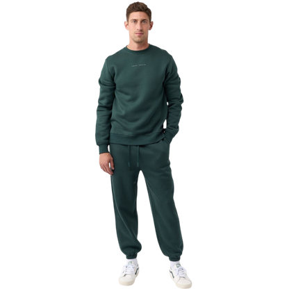 A man with short brown hair is wearing a dark green Men's CityScape Crewneck from Cozy Earth, featuring the words "COLD EARTH" printed on it. He is gazing slightly to the side against a plain white background. |Color:Spruce
