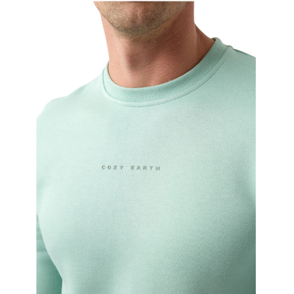 A man with short brown hair is wearing a dark green Men's CityScape Crewneck from Cozy Earth, featuring the words "COLD EARTH" printed on it. He is gazing slightly to the side against a plain white background. |Color:Spruce