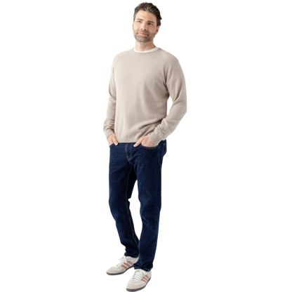 A man stands against a white background wearing a Men's Crewneck Sweater by Cozy Earth, dark blue jeans, and gray sneakers with white and red accents. He has his hands in his pockets and is smiling slightly at the camera. |Color:Sandstone