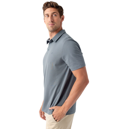 A man with short brown hair, dressed in a light gray Men's Everyday Polo from Cozy Earth and beige pants, poses against a plain white background. He looks slightly to his left, with one hand in his pocket. |Color:Deep Blue