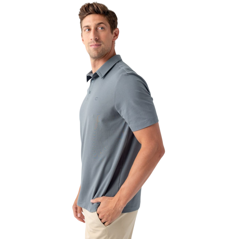 A man with short brown hair, dressed in a light gray Men's Everyday Polo from Cozy Earth and beige pants, poses against a plain white background. He looks slightly to his left, with one hand in his pocket. |Color:Deep Blue