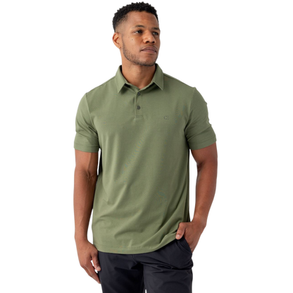 A person stands against a plain background wearing the Men's Everyday Polo by Cozy Earth in green, paired with black pants. They have short hair and are looking off to the side with one hand in their pocket. |Color:Juniper