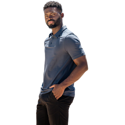 A person with short, light brown hair is wearing the Men's Everyday Polo by Cozy Earth in a light gray shade, paired with light beige pants. They are standing and slightly looking over their left shoulder with a neutral expression against a plain white background. |Color:Stone