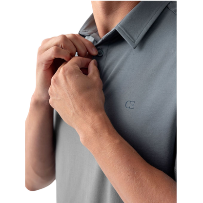 A person with short, light brown hair is wearing the Men's Everyday Polo by Cozy Earth in a light gray shade, paired with light beige pants. They are standing and slightly looking over their left shoulder with a neutral expression against a plain white background. |Color:Stone