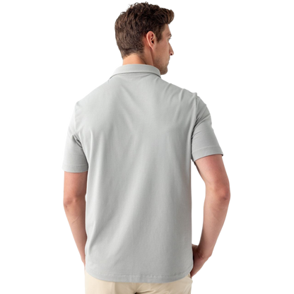 A person with short, light brown hair is wearing the Men's Everyday Polo by Cozy Earth in a light gray shade, paired with light beige pants. They are standing and slightly looking over their left shoulder with a neutral expression against a plain white background. |Color:Stone