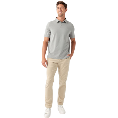 A person with short, light brown hair is wearing the Men's Everyday Polo by Cozy Earth in a light gray shade, paired with light beige pants. They are standing and slightly looking over their left shoulder with a neutral expression against a plain white background. |Color:Stone