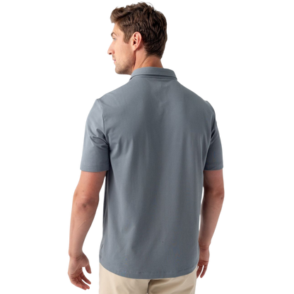 A person with short, light brown hair is wearing the Men's Everyday Polo by Cozy Earth in a light gray shade, paired with light beige pants. They are standing and slightly looking over their left shoulder with a neutral expression against a plain white background. |Color:Stone