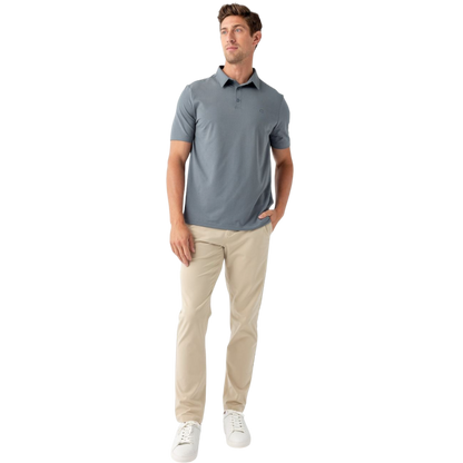 A person with short, light brown hair is wearing the Men's Everyday Polo by Cozy Earth in a light gray shade, paired with light beige pants. They are standing and slightly looking over their left shoulder with a neutral expression against a plain white background. |Color:Stone