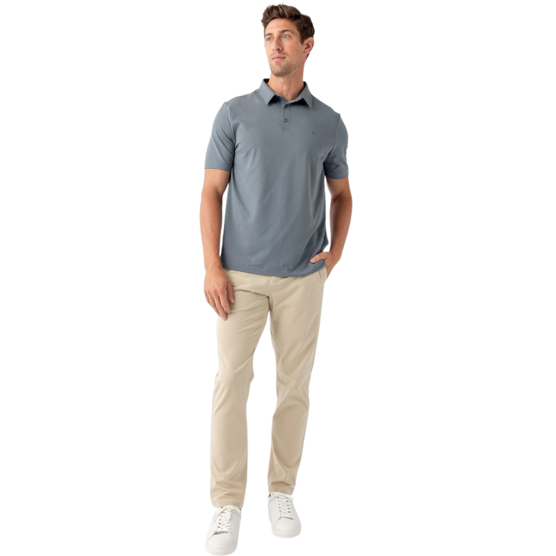 A person with short, light brown hair is wearing the Men's Everyday Polo by Cozy Earth in a light gray shade, paired with light beige pants. They are standing and slightly looking over their left shoulder with a neutral expression against a plain white background. |Color:Stone