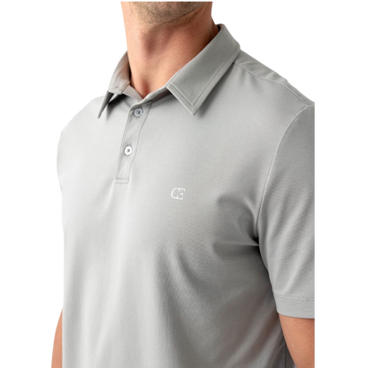 A person with short, light brown hair is wearing the Men's Everyday Polo by Cozy Earth in a light gray shade, paired with light beige pants. They are standing and slightly looking over their left shoulder with a neutral expression against a plain white background. |Color:Stone