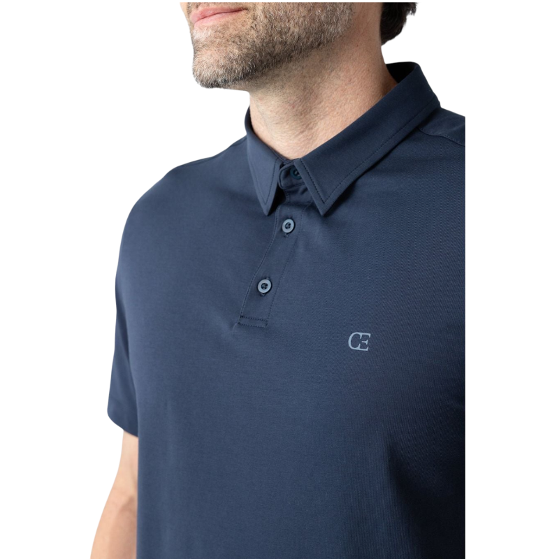 A person with short, light brown hair is wearing the Men's Everyday Polo by Cozy Earth in a light gray shade, paired with light beige pants. They are standing and slightly looking over their left shoulder with a neutral expression against a plain white background. |Color:Stone