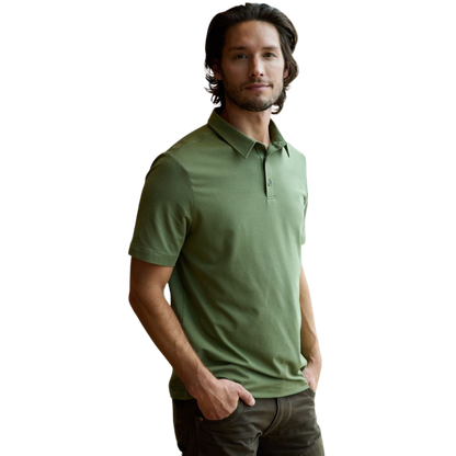 A person with short, light brown hair is wearing the Men's Everyday Polo by Cozy Earth in a light gray shade, paired with light beige pants. They are standing and slightly looking over their left shoulder with a neutral expression against a plain white background. |Color:Stone